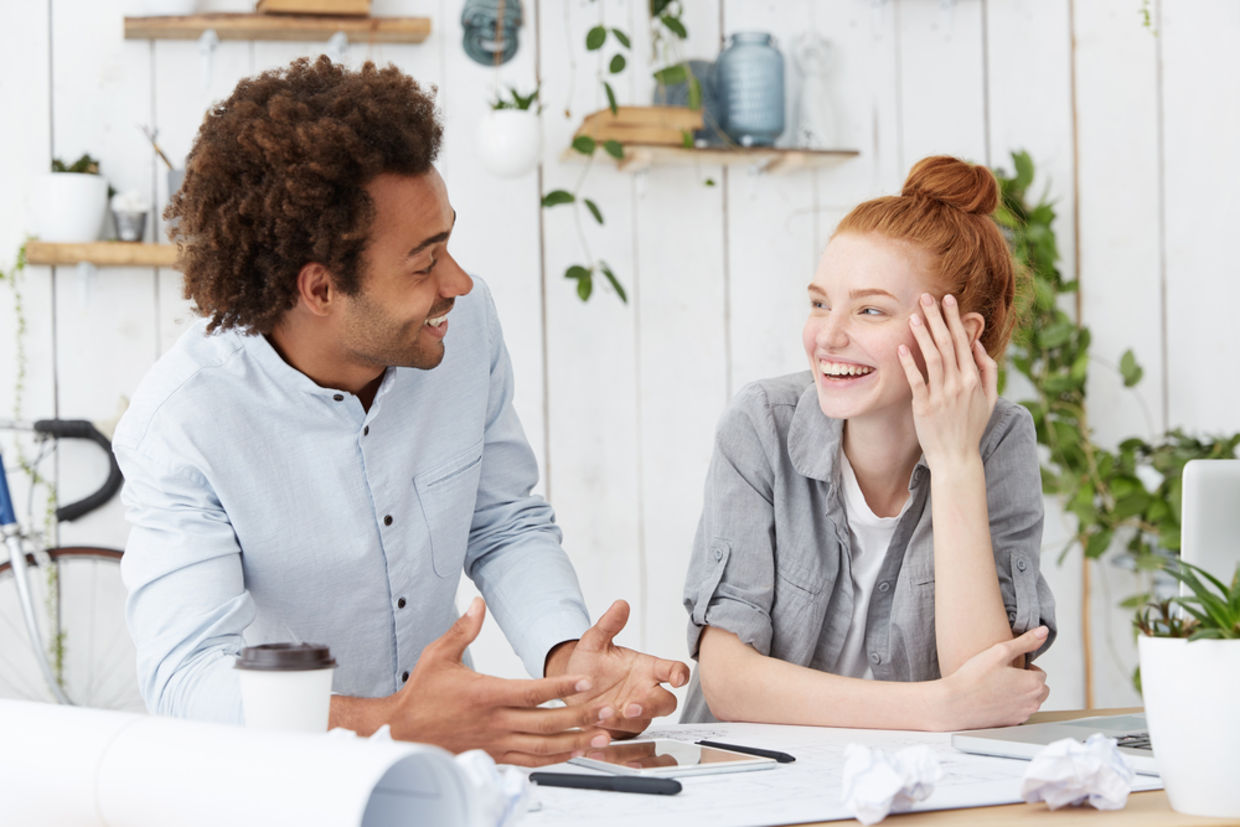 Beautiful young redhead European woman designer with hair bun laughing at jokes of her cheerful charismatic dark-skinned male colleague Beautiful young redhead European woman designer with hair bun laughing at jokes of her cheerful charismatic dark-skinned male colleague