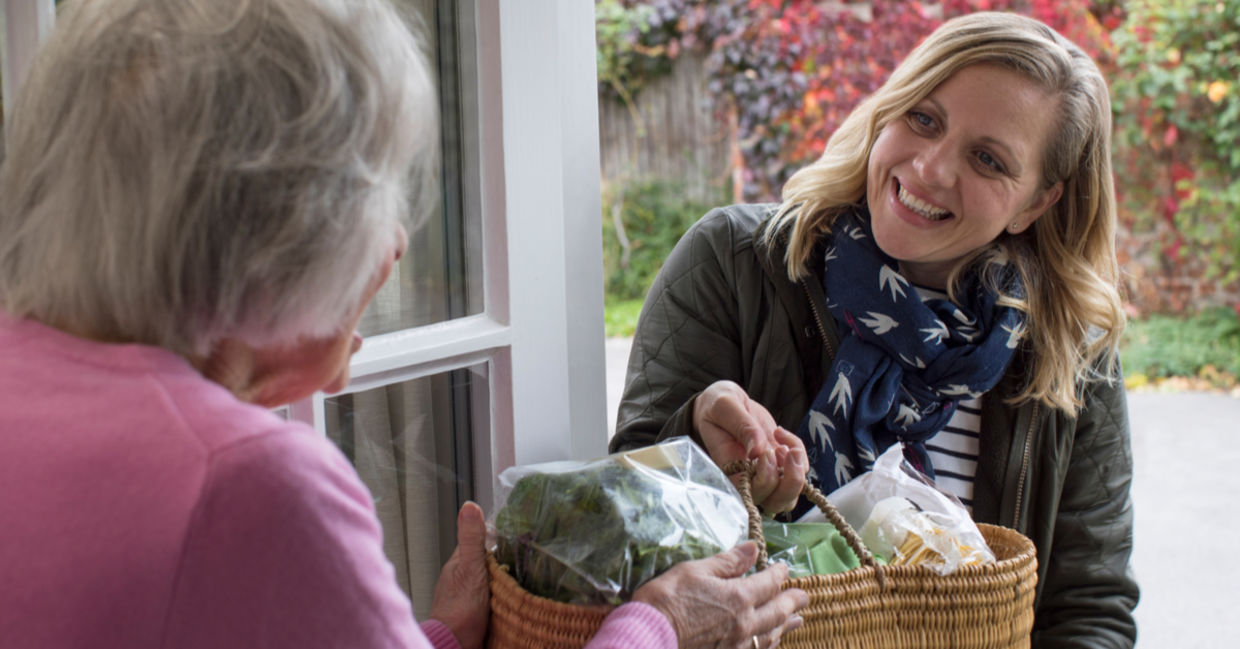 A smiling woman reaches out by delivering a basket of food to a senior. A smiling woman reaches out by delivering a basket of food to a senior.