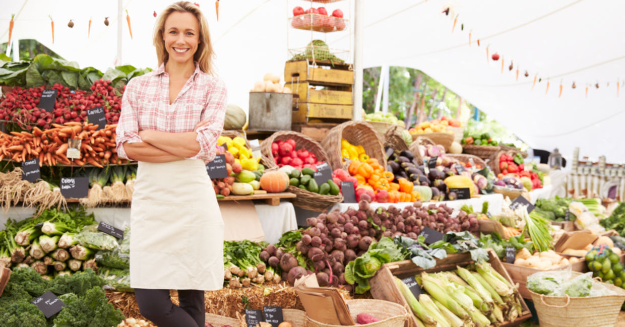 A vendor stands in front of her vegetable stand at an outdoor market. A vendor stands in front of her vegetable stand at an outdoor market.