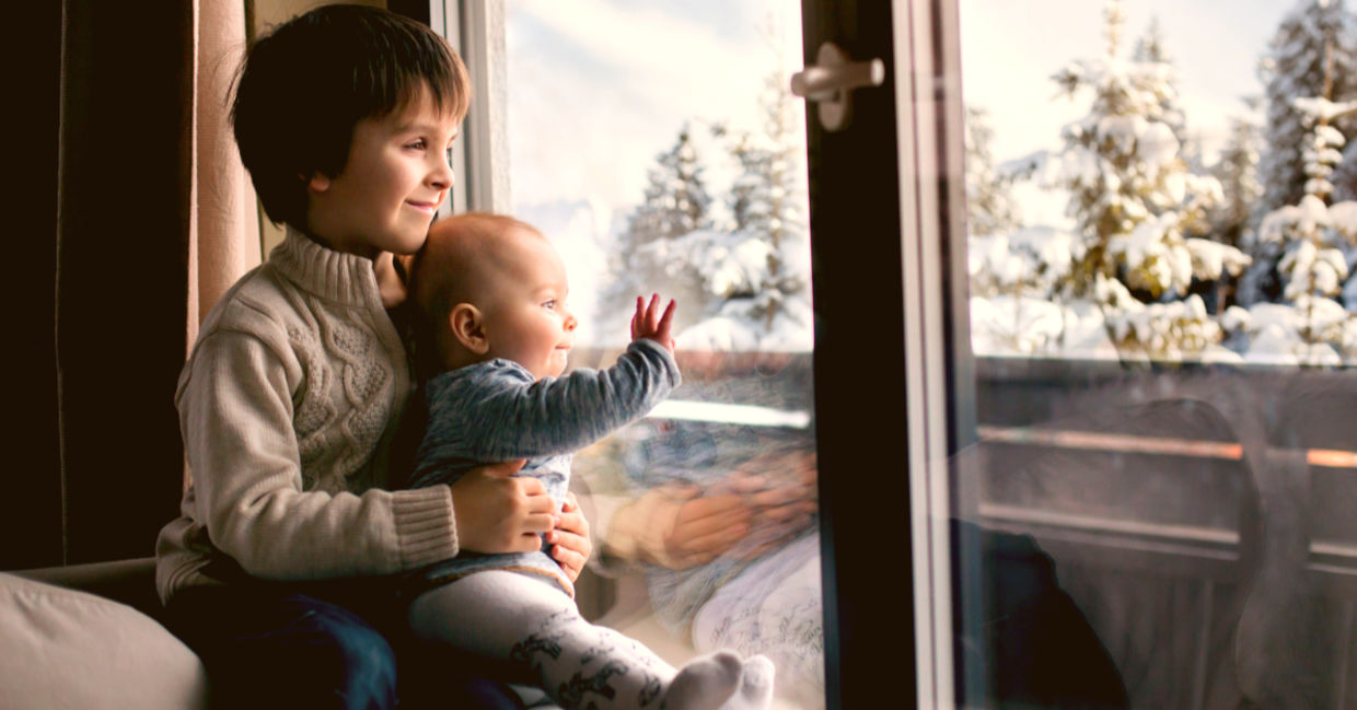Boy holding his baby brother, sitting by the window in living room, looking at a snowy landscape outdoors. Boy holding his baby brother, sitting by the window in living room, looking at a snowy landscape outdoors.