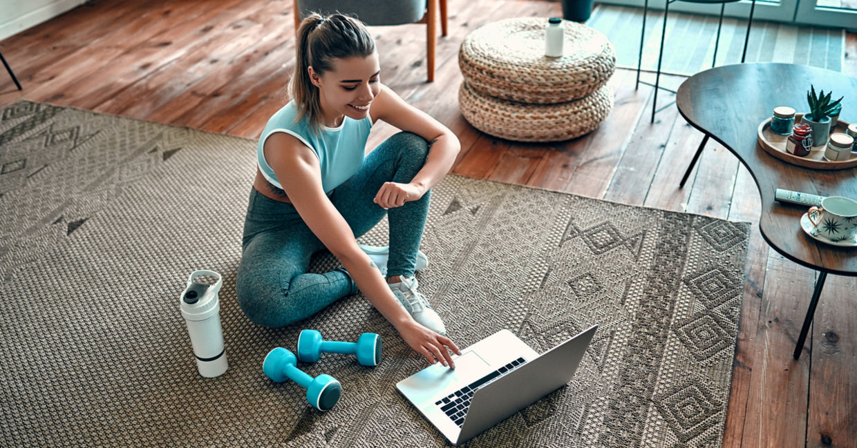 Sporty woman sitting on the floor with sports equipment around her as she uses a laptop. Sporty woman sitting on the floor with sports equipment around her as she uses a laptop.