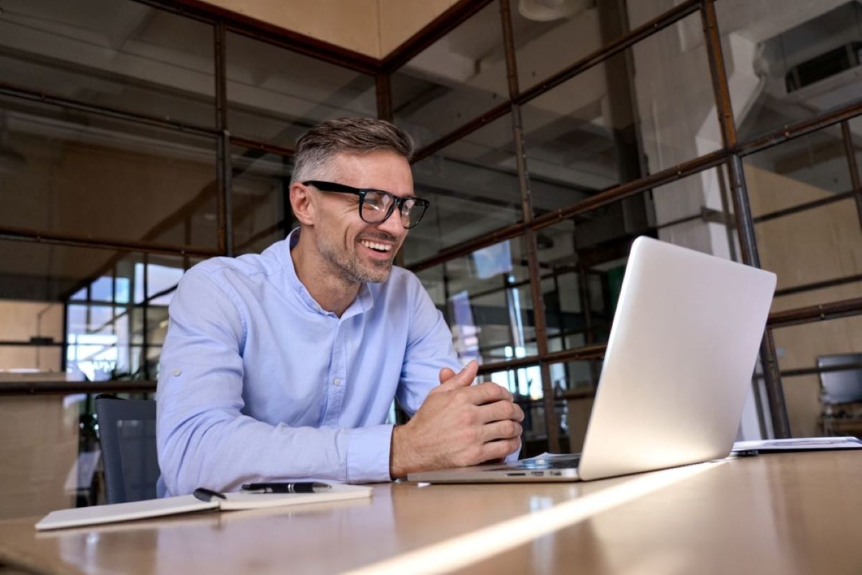 A man wearing glasses smiles while looking at a laptop screen. A man wearing glasses smiles while looking at a laptop screen.